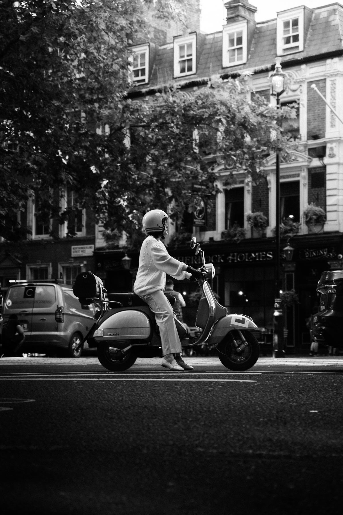Black and white street photograph of a rider on a Vespa stopped outside a London pub with plane trees overhead